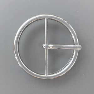 Pode incluir: Fivela de cinto prateada brilhante com design circular. A fivela apresenta uma moldura redonda com uma barra horizontal e uma barra vertical. O metal tem um acabamento brilhante, que reflete a luz. Design simples e elegante.
