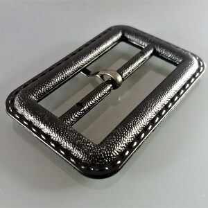 May include: A black metal belt buckle with a textured finish and a stitched edge.
