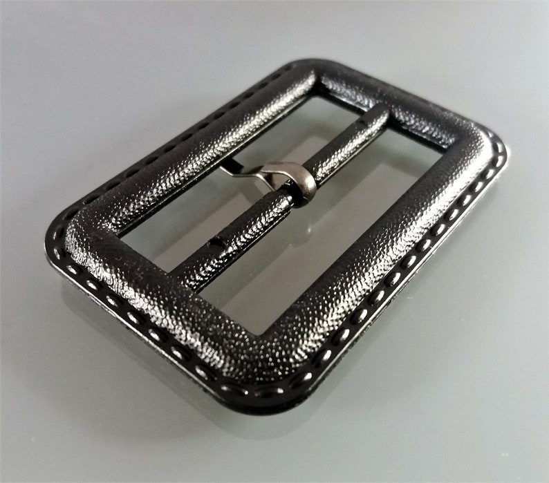 May include: A black metal belt buckle with a textured finish and a stitched edge.