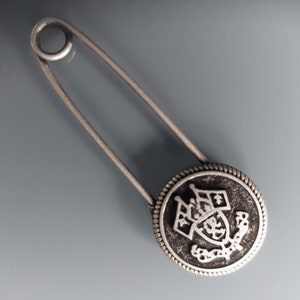 Peut inclure: Une épingle à nourrice en argent avec un bouton rond noir orné d'un blason argenté avec une couronne et le texte "Royal" en cercle.