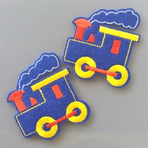Peut inclure: Deux appliques de train en feutre bleu avec des roues jaunes, des cheminées rouges et des détails de fumée blanche.