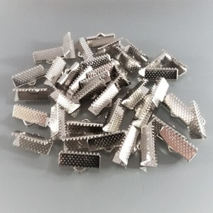 May include: A pile of silver metal clasps for straps or cords. The clasps are rectangular with a textured surface and a small hole on each side.