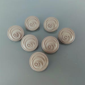 Könnte beinhalten: Sieben taupefarbene Knöpfe mit einem Spiraldesign. Die runden Knöpfe haben ein erhabenes, konzentrisches Wirbelmuster, das von einem zentralen Punkt ausgeht. Die Knöpfe sind auf einer grauen Oberfläche angeordnet.