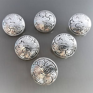 Op de afbeelding: Zes zilverkleurige, ronde knopen met een sierlijk bloemen- en wervelbladontwerp. De knopen hebben een verhoogd, gedetailleerd patroon en een licht gewelfde vorm, geschikt voor naai- of knutselprojecten.