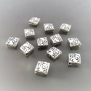 Op de afbeelding: Een verzameling vierkante zilveren kralen met een bloemmotief in zwart. Elke kraal heeft aan elke kant een klein gaatje. De kralen zijn verspreid over een grijze ondergrond, sommige scherp en andere iets wazig.