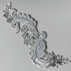Può includere: Applicazione ricamata argentata con design floreale e a volute. L'applicazione presenta cuciture dettagliate, paillettes e un motivo ornato. Ideale per abbellire capi d'abbigliamento o progetti artigianali.