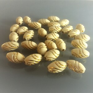 Peut inclure: Un ensemble de perles ovales ivoire, ornées de motifs dorés en lignes courbes. Ces perles, idéales pour le bricolage, sont disposées sur une surface gris clair, mettant en valeur leur design décoratif et leur potentiel en bijouterie.