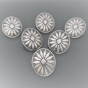 Può includere: Sei bottoni in metallo color argento con un design geometrico. Ogni bottone presenta un quadrato centrale circondato da linee radiali.