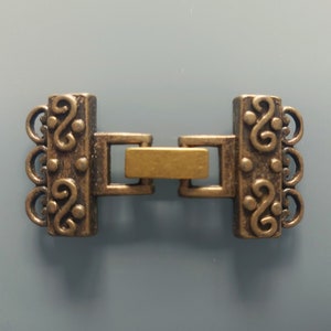 Può includere: Un fermaglio in metallo dorato con un design decorativo. Il fermaglio ha quattro anelli su un lato e una barra scorrevole sull'altro.