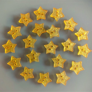 Pode incluir: Uma coleção de 20 botões em forma de estrela dourada com glitter. Os botões estão dispostos em círculo sobre um fundo cinza.