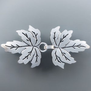 Könnte beinhalten: Silberfarbener Metallverschluss mit einem Blattdesign. Der Verschluss besteht aus zwei Blättern, die durch einen Ring in der Mitte verbunden sind.