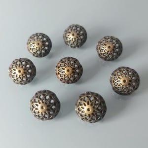 May include: Nine bronze-colored filigree beads with a floral design are arranged on a light gray surface. Each bead has a central gold-colored accent. The beads are spherical and have an open, lacy pattern.