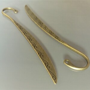 May include: Two antique gold-toned metal bookmarks with a decorative swirl design. The bookmarks have a pointed tip and a hook at the top.