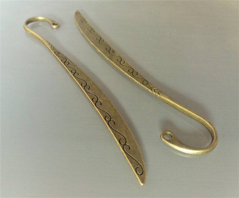 May include: Two antique gold-toned metal bookmarks with a decorative swirl design. The bookmarks have a pointed tip and a hook at the top.