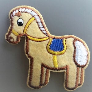 Peut inclure: Une applique en feutre beige en forme de cheval avec des détails brodés en brun, une selle jaune avec des finitions bleues et une queue blanche.