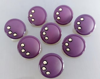 May include: Eight round, purple buttons with three small, silver-colored metal accents arranged in a triangle on each button. The buttons are arranged in a circular pattern against a light gray background.