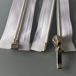 May include: Four white zipper tapes with silver metal teeth and sliders. The zippers are closed and the sliders are at the top of the tapes.