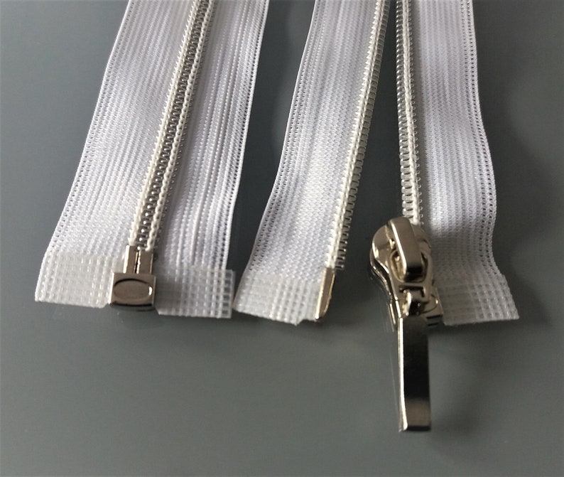May include: Four white zipper tapes with silver metal teeth and sliders. The zippers are closed and the sliders are at the top of the tapes.