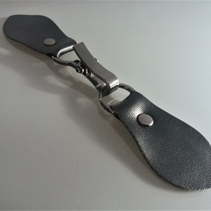 Könnte beinhalten: Ein schwarzer Leder-Toggle-Verschluss mit einer silbernen Metallschlaufe und zwei schwarzen Lederlaschen. Der Verschluss wird zum Verschließen von Kleidung verwendet.