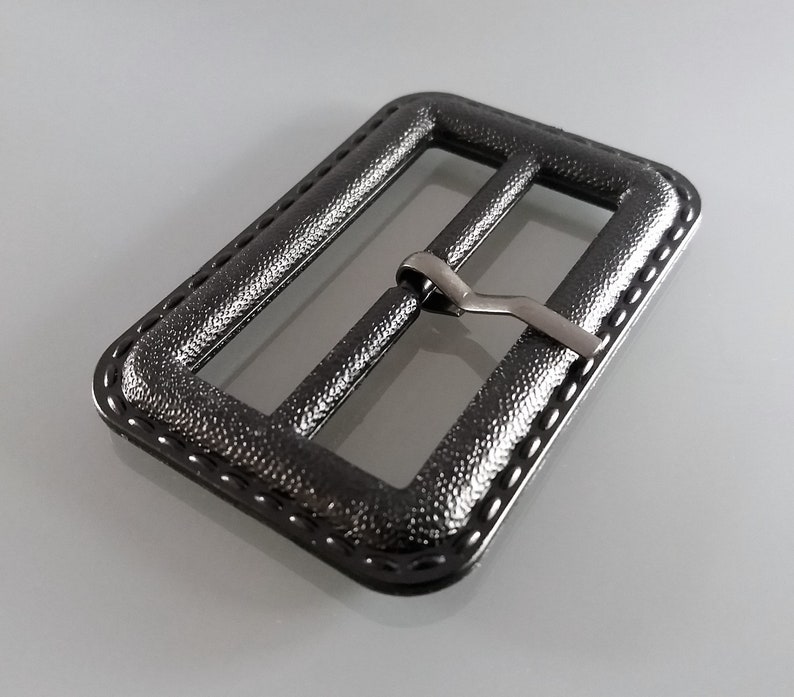 May include: A black metal buckle with a textured finish and a silver metal slider. The buckle has a stitched edge.