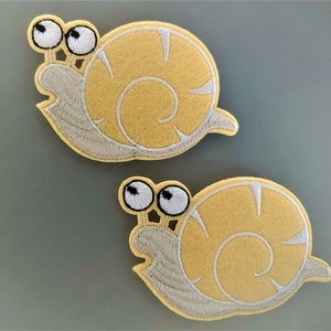 Peut inclure: Deux appliques de snail en feutre jaune avec des yeux blancs et un contour blanc. Les escargots sont tournés dans des directions différentes.