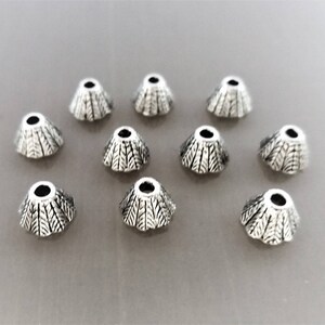 Peut inclure: Dix capuchons de perles argentés en forme de cône. Chaque capuchon présente un motif texturé détaillé et un trou central. Les capuchons sont disposés sur une surface gris clair, parfaits pour la création de bijoux.