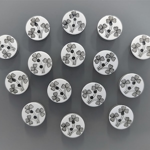 Peut inclure: Un ensemble de 15 boutons blancs avec des motifs de trèfles noirs. Les boutons sont disposés en cercle sur une surface grise.