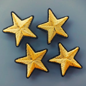 Può includere: Quattro toppe a forma di stella dorate con contorni neri. Le stelle sono ricamate con filo dorato e hanno una texture leggermente in rilievo.
