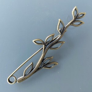 May include: A bronze-coloured brooch shaped like a leafy branch. The pin features a safety clasp and intricate cut-out leaf details. The brooch is set against a grey background.