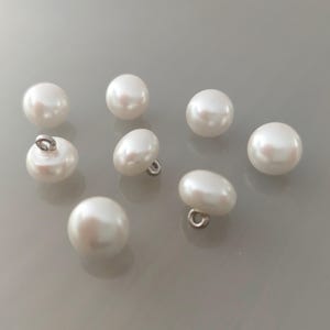 Può includere: Una collezione di otto bottoni bianchi perlati. Alcuni bottoni hanno un anello sul retro per cucire. I bottoni sono rotondi e hanno una finitura liscia e lucida. I bottoni sono sparsi su una superficie grigio chiaro.