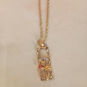May include: Gold-toned necklace featuring a rectangular pendant with a mushroom design. The pendant showcases three colorful mushrooms: one red, one yellow, and one with a crystal-like top. The chain is a delicate gold color.