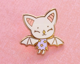 Peut inclure: Une épingle en émail blanche représentant une chauve-souris de dessin animé avec des oreilles roses et un t-shirt à motifs d'étoiles violettes et dorées. La chauve-souris vole avec ses ailes déployées.