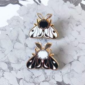 May include: Two black and white enamel pin badges featuring a stylised moth design. The moths have a black and white pattern on their wings and a black or white circle in the centre of their bodies.