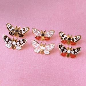 May include: Six gold-tone enamel pins shaped like moths with black, white and pink patterns. The moths have a detailed design with a cloud-like pattern on their wings.