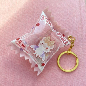 May include: A pink and white candy-shaped keychain with a clear plastic window. Inside the window is a small white creature with wings and a pink and purple design. The keychain has a gold metal ring and chain.