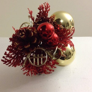 May include: A festive Christmas ornament arrangement featuring gold and red baubles, pinecones, and decorative red sprigs. The arrangement includes miniature brass French horns, creating a unique holiday decoration.