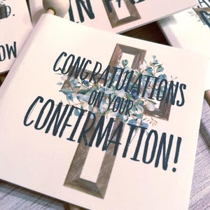 May include: White card with a wooden cross design and the text "CONGRATULATIONS ON YOUR CONFIRMATION!". The cross is decorated with blue and green floral accents. Other cards with similar designs are visible in the background.