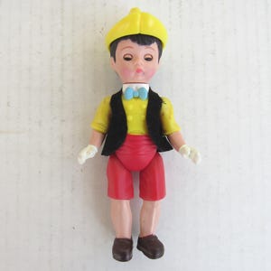 May include: A vintage doll of a young boy, wearing a yellow hat, shirt, and white gloves. He has a black vest, red shorts, a blue bow tie, and brown shoes. The doll is standing against a white background.