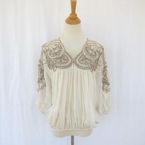 Puede incluir: Blusa color crema con intrincados detalles bordados en un tono neutro alrededor del escote y las mangas. La blusa presenta un corpi&ntilde;o fruncido y mangas tres cuartos, exhibida en un maniqu&iacute; sobre un fondo blanco.