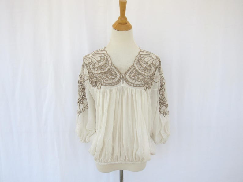 Puede incluir: Blusa color crema con intrincados detalles bordados en un tono neutro alrededor del escote y las mangas. La blusa presenta un corpi&ntilde;o fruncido y mangas tres cuartos, exhibida en un maniqu&iacute; sobre un fondo blanco.