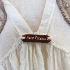 Puede incluir: Primer plano de una blusa color crema con dise&ntilde;o de cuello en V. Una peque&ntilde;a etiqueta rectangular de color cobre con las palabras "Free People" est&aacute; centrada en el escote. La blusa tiene un delicado ribete texturizado y un detalle de ganchillo.