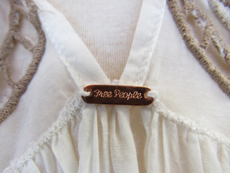 Puede incluir: Primer plano de una blusa color crema con dise&ntilde;o de cuello en V. Una peque&ntilde;a etiqueta rectangular de color cobre con las palabras "Free People" est&aacute; centrada en el escote. La blusa tiene un delicado ribete texturizado y un detalle de ganchillo.