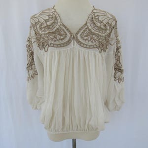 Puede incluir: Blusa color crema con intrincados bordados marrones alrededor del escote y las mangas. La blusa presenta un cuello en V y detalles de tela fruncida, creando una silueta fluida. Las mangas son de tres cuartos.