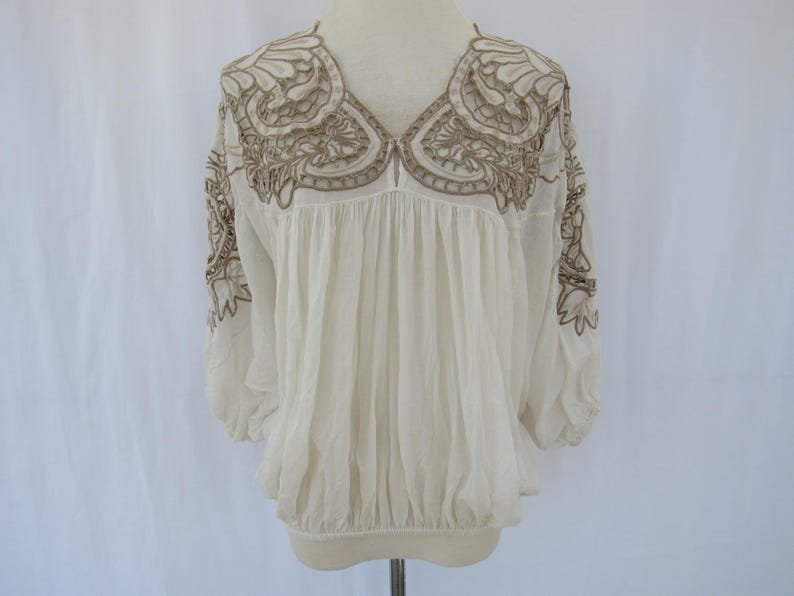 Puede incluir: Blusa color crema con intrincados bordados marrones alrededor del escote y las mangas. La blusa presenta un cuello en V y detalles de tela fruncida, creando una silueta fluida. Las mangas son de tres cuartos.