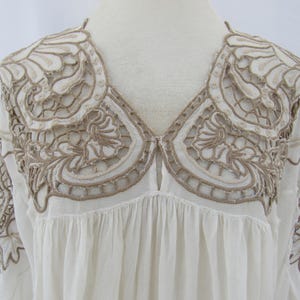 Puede incluir: Blusa color crema con intrincados bordados calados en un tono marr&oacute;n contrastante. El bordado adorna los hombros y el escote, creando un canes&uacute; decorativo. La blusa tiene cuello en V y un cuerpo fruncido y fluido, lo que sugiere un estilo bohemio.