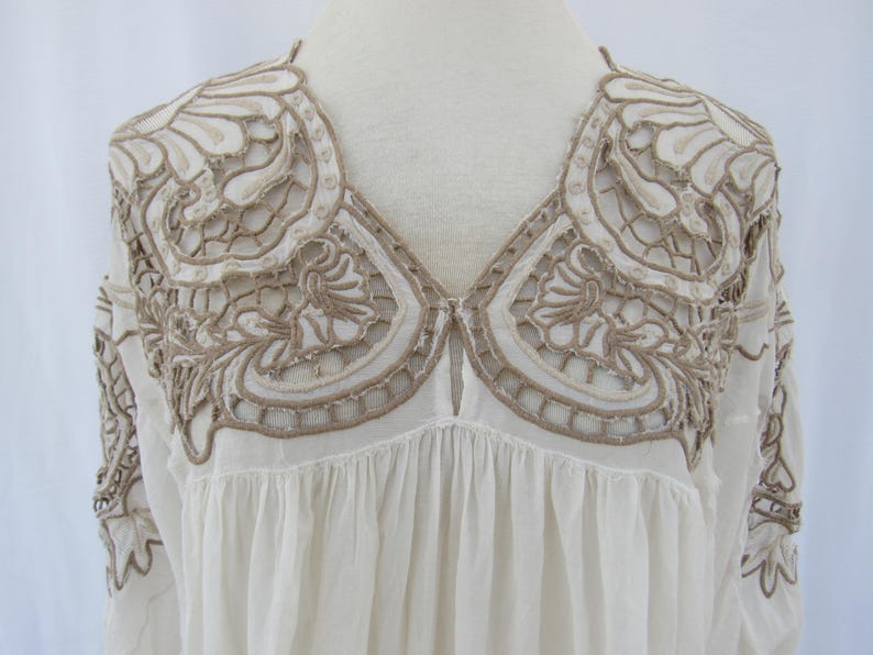 Puede incluir: Blusa color crema con intrincados bordados calados en un tono marr&oacute;n contrastante. El bordado adorna los hombros y el escote, creando un canes&uacute; decorativo. La blusa tiene cuello en V y un cuerpo fruncido y fluido, lo que sugiere un estilo bohemio.