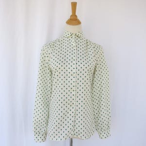 May include: A long-sleeved, button-down blouse on a wooden mannequin. The blouse is cream-colored with a repeating pattern of small, dark green heart shapes. The collar is pointed, and the sleeves are cuffed.