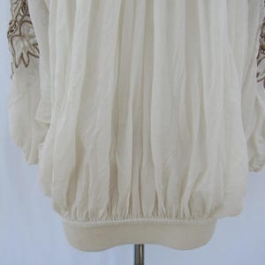 Puede incluir: Blusa color crema con mangas largas y dobladillo el&aacute;stico. Las mangas presentan intrincados patrones florales bordados en un color marr&oacute;n contrastante. La tela parece ligera y ligeramente transparente, con un dise&ntilde;o fruncido.