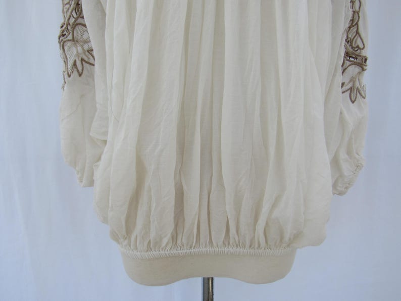 Puede incluir: Blusa color crema con mangas largas y dobladillo el&aacute;stico. Las mangas presentan intrincados patrones florales bordados en un color marr&oacute;n contrastante. La tela parece ligera y ligeramente transparente, con un dise&ntilde;o fruncido.