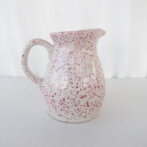 May include: A white ceramic pitcher with a rounded body, a curved handle, and a pouring spout. The pitcher is speckled with small, irregular red dots. The pitcher is likely used for serving beverages.